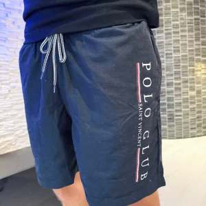 Svarta shorts Polo Club Saint Vincent - Snygga svarta shorts från Polo Club Saint Vincent med vit snörning i midjan och tryckt logga längs ena benet. Shortsen har meshfoder på insidan och är perfekta för sommaren. Klassisk och sportig look med normal passform.