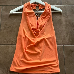 Orange ärmlös halterneck topp - Säljer en orange/korall halterneck topp med snygg drapering framtill och öppen rygg med ringdetalj och smala band. Perfekt för dig som vill sticka ut med en unik och trendig look. Den har ett litet hål på framsidan som sytts ihop sen tidigare, detta syns inte mycket när man har på sig den. Storlek ca S, passar allt från XS-M. 