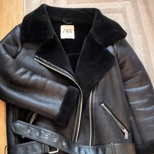 Svart pälsjacka från Zara med bälte - Använt få få gånger Säljer en svart pälsjacka från Zara med bred krage, mjuk insida och detaljer som dragkedjor och bälte i midjan. Jackan har långa ärmar och en cool bikerstil som passar perfekt när du vill ha en edgy look. Fodret är fluffigt och värmande.