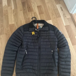 Svart pufferjacka från Parajumpers - Säljer en svart pufferjacka från Parajumpers med klassisk patch på ärmen och gul detalj vid kragen. Jackan har dragkedja framtill, två fickor med dragkedja och hög krage. Perfekt för kyliga dagar och riktigt snygg till streetwear.