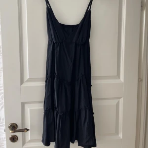 Svart långklänning med volanger - Svart långklänning med smala axelband och v-ringad urringning. Klänningen har flera volanglager som ger en flowy och avslappnad look. Perfekt för varma dagar och enkel att styla med accessoarer. Materialet känns mjukt och lätt.