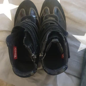 Svarta sneakers från Tamaris med dragkedja - Snygga svarta sneakers från Tamaris med både dragkedja och kardborreband. Skorna har en blandning av blankt syntetmaterial och mockadetaljer, samt vit sula och dekorativa sömmar. Perfekta för dig som gillar en sportig och cool stil. Använda några gånger. 25.5 cm i innersula 16-17 cm höga.