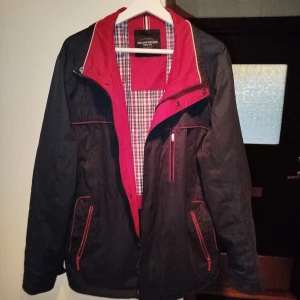 vindjacka från Erla of Sweden storlek C50/m-l - Svart vindjacka med röda detaljer och dragkedjor från Erla of Sweden. Jackan har röd insida och rutigt foder i rött, blått och vitt. Flera fickor med dragkedja och snygga kontrastsömmar. Perfekt för dig som vill ha en stilren och sportig look.