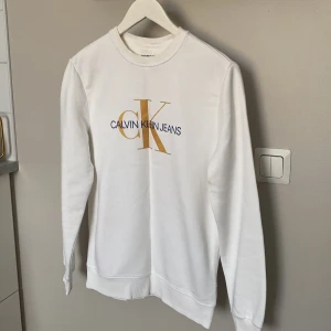 Vit sweatshirt från Calvin Klein Jeans - Vit sweatshirt från Calvin Klein Jeans med stort guldigt och blått logotryck på bröstet. Klassisk rund halsringning och långa ärmar. Mjuk och skön i bomullsmix, perfekt för en clean och stilren look.
