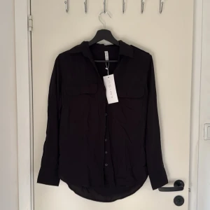 Svart skjorta - Svart skjorta från New Yorker i storlek XS. Skjortan har klassisk krage, knäppning framtill, två bröstfickor med lock och axelklaffar. Materialet känns lätt och smidigt, perfekt till många olika outfits. Endast testad, prislappen kvar☺️