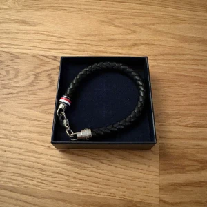 Tommy Hilfiger armband  - Stilrent svart flätat armband från Tommy Hilfiger. Snyggt och enkelt att matcha till din outfit. Levereras i originalask med logga. Bara använd en gång. Helt ny