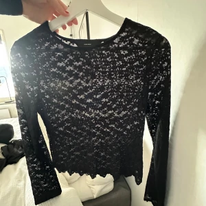 Svart spetsblus från Vero Moda - Säljer en svart långärmad blus från Vero Moda i transparent spets med blommigt mönster. Toppen har rund halsringning och vågig kant nedtill för en extra snygg touch. Perfekt att styla med jeans eller kjol för en cool look.