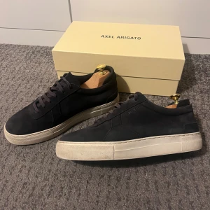 Blå arigato skor  - Säljer ett par snygga axelarigato i mocka. På bilden ser de svarta ut men de är mörkblåa egentligen. Skickat är bra då skorna har tagits väl hand om under användning. Nackdel är att en bit gummi har lossnat som man kan se på bild 4. Box och arigato påse medföljer såklart. Storlek 43. Hör av er vid frågor! 