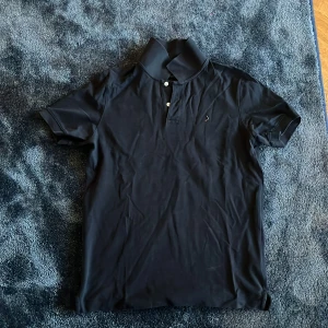 Svart pikétröja från Boomerang - Säljer en svart pikétröja från Boomerang i klassisk modell. Tröjan har korta ärmar, krage och knäppning med två knappar framtill. Tillverkad i 100% ekologisk bomull och har en liten broderad logga på bröstet. Perfekt för en clean och stilren look.