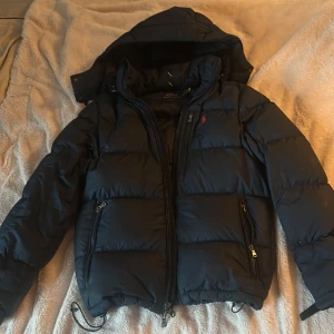 Marinblå pufferjacka från Polo Ralph Lauren - Säljer en marinblå pufferjacka från Polo Ralph Lauren med huva och dragkedja framtill. Jackan har två fickor med dragkedja och den klassiska röda Polo-loggan på bröstet. Perfekt för kalla dagar och riktigt snygg passform.