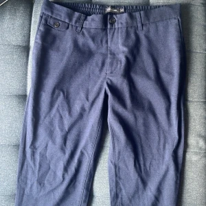 Mörkblå kostymbyxor Dressmann Slim - Mörkblå kostymbyxor från Dressmann i slim fit-modell. Byxorna har klassiska fickor med knappdetaljer och bälteshällor. Perfekta för en clean och stilren look. Materialet känns mjukt och har en lätt glans.