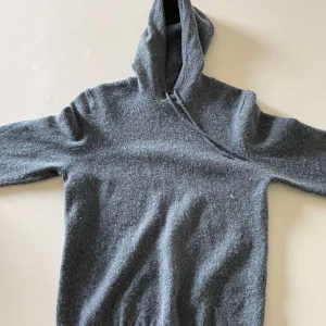 Grå stickad hoodie med huva - Mysig grå stickad hoodie med huva och långa ärmar. Tröjan har en enkel design utan synliga loggor eller mönster och är perfekt för kyliga dagar. Ribbad nederkant ger en skön passform och den stickade ullblandningen gör den extra varm.