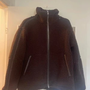 Brun teddyjacka från H&M  - Mysig brun teddyjacka från H&M med hög krage och hel dragkedja framtill. Jackan har två snedställda fickor med kant i matchande färg och är tillverkad i ett fluffigt teddy-material som håller dig varm. 