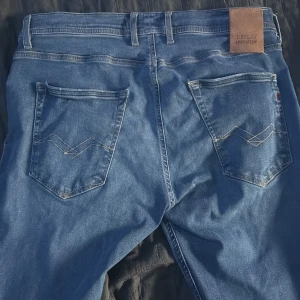 Blå Replay Grover Hyperflex jeans - Säljer ett par blå Replay Grover Hyperflex jeans med klassisk femficksdesign och snygga kontrastsömmar på bakfickorna. Jeansen har normal passform och är tillverkade i stretchigt jeansmaterial för extra komfort. Perfekta för dig som gillar stilrena och bekväma jeans.