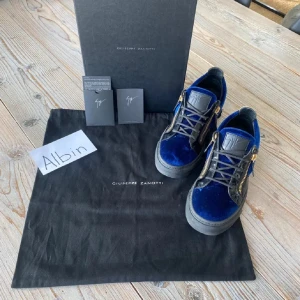 Blå sneakers från Giuseppe Zanotti - Säljer ett par riktigt snygga sneakers från Giuseppe Zanotti i blå sammet med svarta lackdetaljer och guldiga dragkedjor på sidorna. Skorna har svarta snören och en rejäl svart sula. Perfekta för dig som vill sticka ut med exklusiv stil. Storlek är 40 och skicket är 9:10