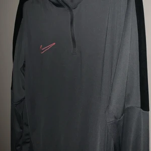 Svart Nike Dri-Fit hoodie half zip - Svart hoodie från Nike med röd broderad swoosh på bröstet. Tröjan är i Dri-Fit-material som andas och har en half zip framtill. Långärmad och perfekt för träning eller chill. Snygg och sportig look.
