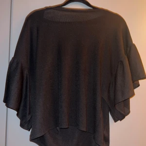 Svart topp med volangärm från Zara - En svart topp från Zara med vida volangärmar och rund halsringning. Tunt, lätt ribbat material, det är storlek S