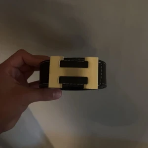 Svart bälte med H - Snyggt svart bälte i skinn med ikoniskt H-format guldspänne. Bältet har vita kontrastsömmar längs kanterna och är brett, vilket ger en riktigt lyxig och stilren känsla. Perfekt accessoar för att lyfta vilken outfit som helst. Man kan ta bort H:et och bältet är 105 cm långt