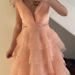 Ljusrosa volangklänning med paljetter - Superfin balklänning i ljusrosa/aprikos med flera lager volanger och ett glittrigt paljettbälte i midjan. Perfekt för bal eller student! Materialet känns lätt men är väldigt fylligt och tjockt då det är mycket lager vilket ger den puffiga looken. Skriv gärna till mig för fler o tydligare bilder (Pris kan diskuteras)