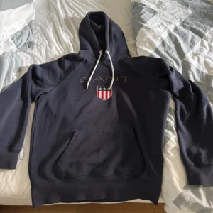 Mörkblå hoodie från GANT med tryck - Säljer en mörkblå hoodie från GANT med vit snörning i huvan och klassiskt GANT-tryck samt sköld på bröstet. Tröjan har känguruficka framtill och är tillverkad i mjuk bomull. Perfekt för en avslappnad och stilren look.