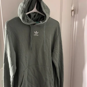 Grön hoodie från Adidas med huva - Säljer en grön hoodie från Adidas med klassisk logga på bröstet. Tröjan har huva med snörning och en stor magficka. Tillverkad i ett mjukt bomullsmaterial och har långa ärmar. Perfekt för en chill och sportig stil.