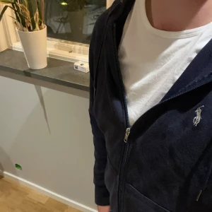 Marinblå hoodie från Polo Ralph Lauren - Säljer en marinblå hoodie från Polo Ralph Lauren med dragkedja och klassisk logga på bröstet. Tröjan har huva med snörning, två fickor fram och är tillverkad i mjuk bomull. Perfekt för chill dagar eller när du vill ha en clean och stilren look.