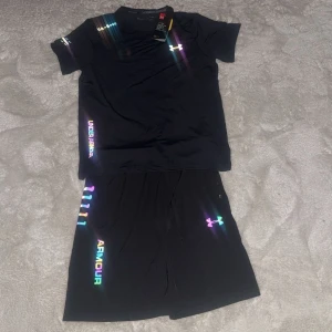 Svarta träningsshorts från Under Armour - Svarta träningsshorts från Under Armour med färgglada loggor och detaljer längs sidan. Shortsen har elastisk midja med snörning och är tillverkade i ett lätt och snabbtorkande syntetmaterial, perfekt för träning. Snygg loose fit och sportig vibe.