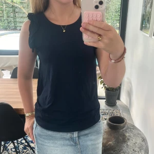 Mörkblå ärmlös topp med volang - Snygg mörkblå ärmlös topp med volangdetaljer på axlarna. Toppen har rund halsringning och en rak passform som är enkel att styla till både jeans och kjol. Perfekt för dig som gillar en stilren look med en twist.