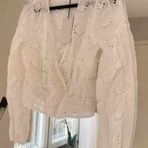 NA-KD blus  - Superfin vit blus med v-ringning och anglaise mönster. Långa ärmar med lätt puff. Blusen har omlottkänsla och är helt ny med prislapp