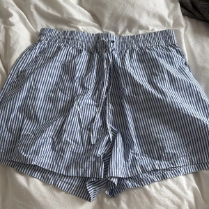 Randiga blåvita shorts med snörning - Säljer ett par luftiga shorts med blå och vita ränder. De har elastisk midja med snörning. Perfekta för varma sommardagar och har en avslappnad passform. Använd som pyjamas shorts! 