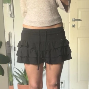 Svart volangkjol med snörning - Supersöt svart kortkjol med flera volanger och snörning i midjan. Kjolen har en loose passform och är tillverkad i mjuk bomull, perfekt för dig som gillar en chill och trendig stil. Enkel att matcha med crop tops eller hoodies.