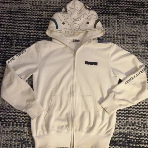 Bape Hoodie - En vit Bape hoodie som jag säljer då den är lite för liten för min smak. Tröjan är använd men ändå i gott skick. Finns en liten fläck av bläck som visas på bild 5, men den syns knappt alls när man har på sig tröjan. Alla svarta detaljer reflekterar även ljus. Som många vet är Bape små i storleken, så den sitter mer som en S även om storleken är M.