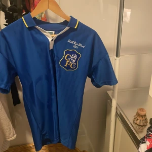 Chelsea retro fotbollströja jubileumströja från FA Cup finalen 1997. - Retro Chelsea FC fotbollströja från Score Draw i klassisk blå färg med gul detalj på kragen. Kortärmad modell med vit knappslå och broderad klubbmärke på bröstet. Materialet är glansigt och lätt, perfekt för dig som gillar vintage-stil. Köpt direkt från Chelseamegastore.com fullt äkta!