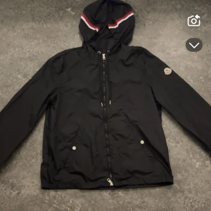 Svart vindjacka från Moncler - Svart vindjacka från Moncler med huva som har röd och vit rand. Jackan har dragkedja framtill, två fickor med tryckknappar och Moncler-logga på ärmen. Perfekt för dig som vill ha en stilren och sportig look.