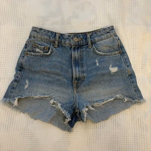 Blå jeansshorts från ZARA, strl 34 - Säljer ett par blå jeansshorts från ZARA i storlek 34. De är högmidjade med fransiga kanter och slitna detaljer framtill. Klassisk femficksmodell med knapp och dragkedja. 