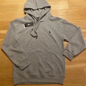Grå hoodie från Polo Ralph Lauren - Snygg grå hoodie från Polo Ralph Lauren med klassisk broderad logga på bröstet. Tröjan har huva med snörning och en stor magficka. Tillverkad i mjuk bomullsmix, perfekt för chill dagar eller när du vill vara casual men ändå stilren.
