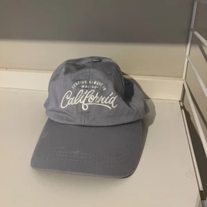 Ljusblå keps med California-brodyr - Snygg ljusblå keps med vit brodyrtext 'California' och 'Surfing always in Malibu' framtill. Klassisk böjd skärm och justerbar rem baktill i metall. Perfekt accessoar för en avslappnad och cool look.