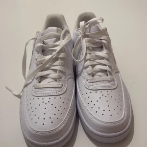 Nike Air Force 1 helvita sneakers - Nike Air Force 1 sneakers i helvitt skinn med klassisk låg siluett. Skorna har perforerad tå, vit sula och snörning. Swoosh-logga på sidan och broderad Nike-text på hälen. Perfekta för en clean och stilren look.