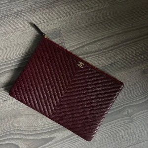 Vinröd kuvertväska  - Säljer en lyxig vinröd kuvertväska i skinn med quiltad struktur och guldigt CC-logo framtill. Väskan har dragkedja och insida i svart satin med märkning. Perfekt för dig som vill ha något stilrent och exklusivt.