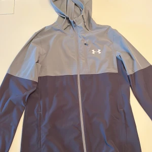 Under Armour grå och mörkblå vindjacka - Snygg tvåfärgad vindjacka från Under Armour i grått och mörkblått. Jackan har huva, hel dragkedja och logga på bröstet. Perfekt för blåsiga dagar och sportiga outfits. Lätt och smidig med praktiska fickor.
