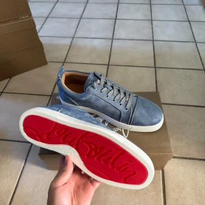 Blå mocka sneakers från Louboutin storlek 45 - Snygga blå sneakers från Christian Louboutin i mjuk mocka med klassisk röd sula och vita kanter. Skorna har rund tå, platt sula och grå snörning. Perfekta för dig som vill sticka ut med exklusiva detaljer och stilren design.
