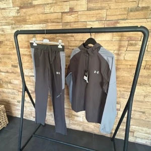 Under Armour grå träningsbyxor - Snygga träningsbyxor från Under Armour i mörkgrått med ljusgrå paneler på sidorna. Byxorna har resår och snörning i midjan, dragkedja vid benslut och reflekterande detaljer. Perfekta för löpning eller gymmet.