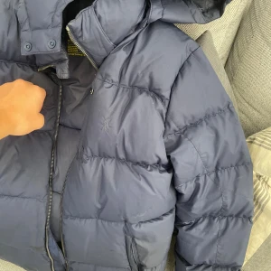 Blå pufferjacka Polo Ralph Lauren XL - Snygg mörkblå pufferjacka från Polo Ralph Lauren med huva och dragkedja. Jackan har quiltad design, två fickor med dragkedja och diskret broderad logga på bröstet. Perfekt för kalla dagar när du vill hålla stilen och värmen. Storlek 170cm. Nypris 3 tusen