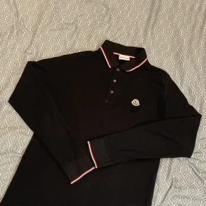 Svart långärmad pikétröja Moncler - Svart långärmad pikétröja från Moncler med klassisk krage och knäppning framtill. Krage och ärmslut har röda och vita ränder. Broderad Moncler-logga på bröstet. Tillverkad i bomull, perfekt för dig som gillar stilrena vibes. Axelbredd 42cm Längd 60cm