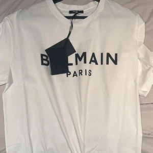 Vit Balmain Paris t-shirt - Vit t-shirt från Balmain Paris med svart logotyptryck över bröstet. Klassisk rund halsringning och korta ärmar. Tillverkad i mjuk bomull för en clean och stilren look. Perfekt för dig som gillar exklusiva streetwear-vibbar.