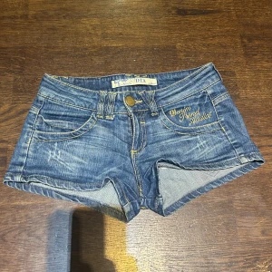 Blå jeansshorts från DTA Addict - Snygga blå jeansshorts från DTA Addict med gul broderad text och cool stjärndetalj på bakfickan. Klassisk femficksmodell, låg midja och slitna detaljer för en chill vibe. Gult kontraststickat och nitar på bakfickan ger extra edge. Perfekt för sommarens häng!