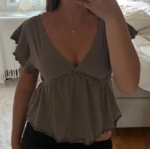 Grå volangblus från 157 - Säljer en grå blus från 157 använd en gång med v-ringad hals och volangdetaljer vid ärmar och nederkant. Blusen har korta, rynkade ärmar och är tillverkad i ett lätt, krinklat material som ger en skön och luftig känsla. Perfekt för dig som gillar en feminin och chill stil.