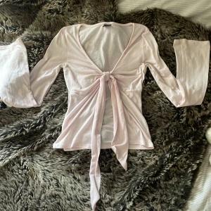 Ljusrosa omlottopp från Gina Tricot - Supersöt ljusrosa omlottopp från Gina Tricot i mjuk bomull. Toppen har långa vida ärmar, v-ringning och knyt framtill som ger en chill och feminin vibe. Perfekt att styla med jeans eller kjol för en trendig look.