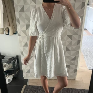 Vit omlottklänning med volang - Säljer en söt vit omlottklänning med volangkant längs v-ringningen och knyt i midjan. Klänningen har korta puffärmar och är tillverkad i ett lätt, strukturerat material som känns luftigt och fräscht. Perfekt för varma dagar och ger en romantisk vibe.