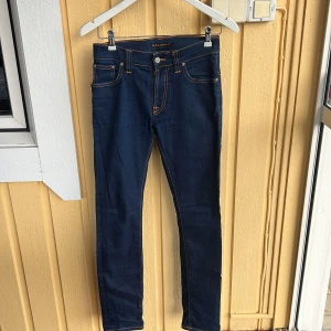 Mörkblå skinny jeans från Nudie Jeans - Snygga mörkblå skinny jeans från Nudie Jeans med klassiska orange sömmar och ikonisk broderad bakficka. Modellen har normal midja och är tillverkad i stretchig bomullsdenim för en tight passform. Perfekta för dig som gillar stilrena och tidlösa jeans.